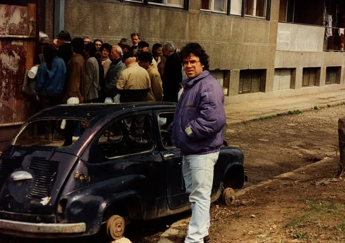 Ο Αργύρης Ντινόπουλος δίπλα σε παλιό αυτοκίνητο, με ουρά ανθρώπων έξω από πολυκατοικία στο βάθος, Σαράγιεβο, 1992.