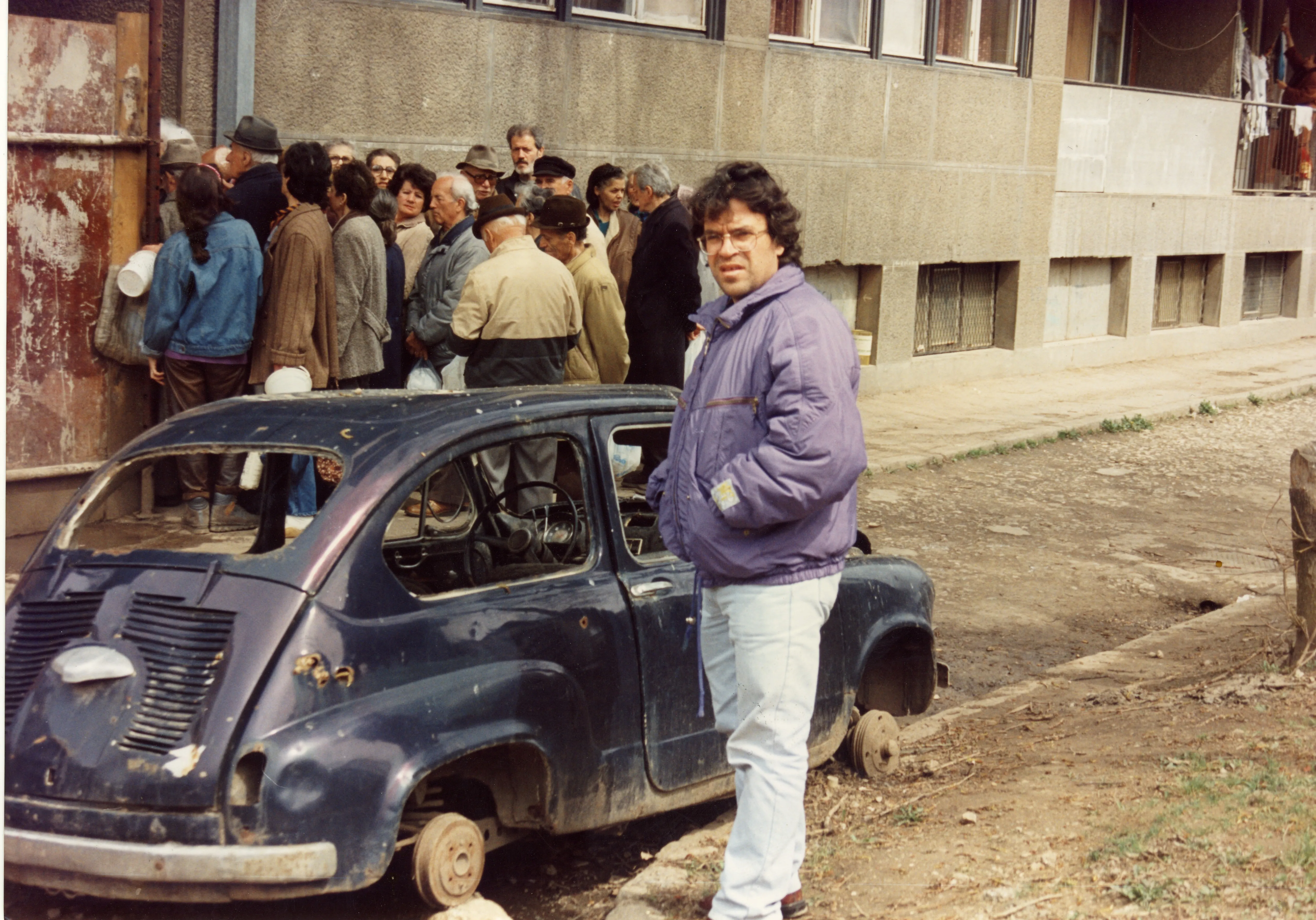 Ο Αργύρης Ντινόπουλος δίπλα σε διαλυμένο αυτοκίνητο χωρίς τροχούς· πίσω, ουρά ανθρώπων με δοχεία κατά μήκος πολυκατοικίας.