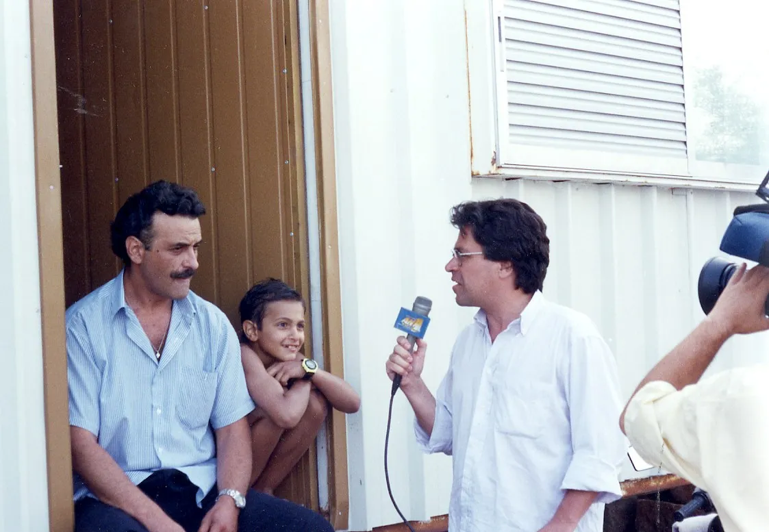 A man with a moustache in a pale blue striped shirt sits in the doorway of a white prefabricated unit; beside him, in the opening of the brown door, a boy, bare-chested, wearing a watch with a yellow face. Facing them a man in a white shirt and glasses holds a microphone with an Antenna flag; at the right edge of the frame a camera operator is visible.