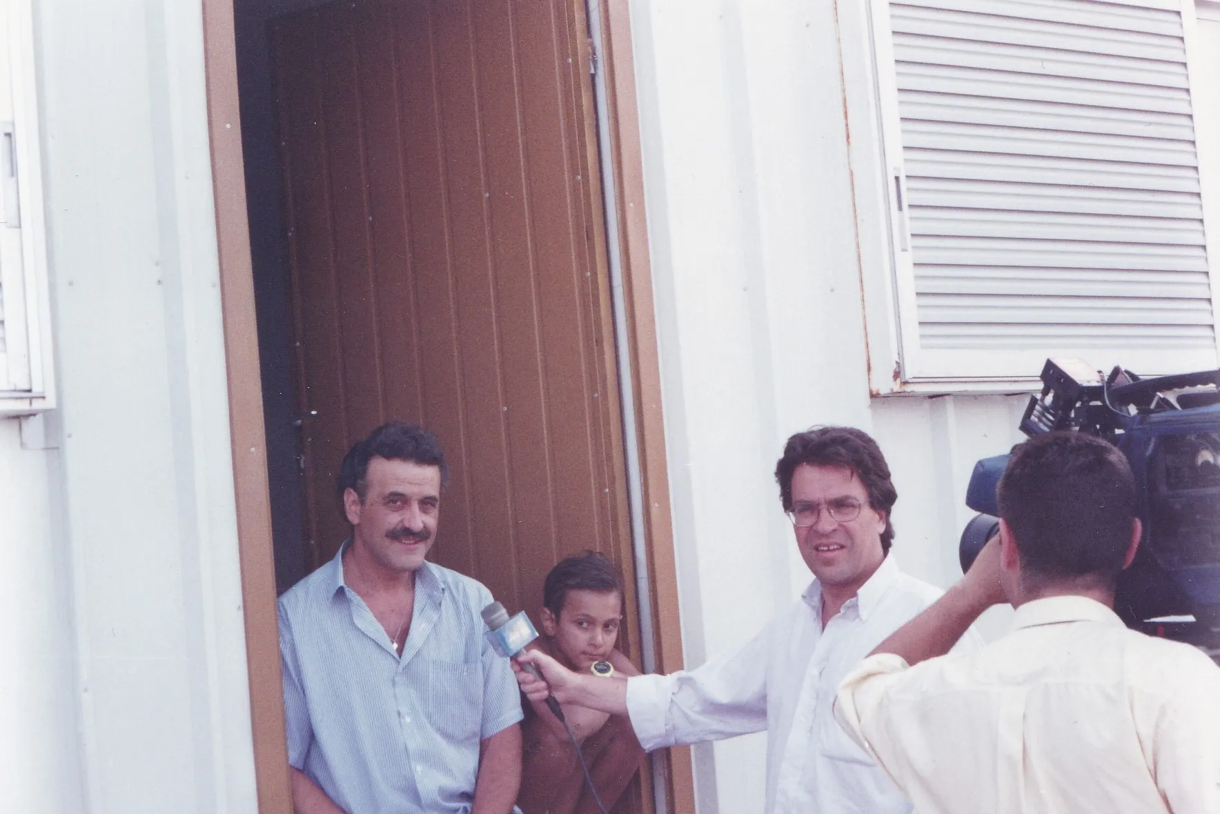 A wider view of the same doorway. The man in the pale blue striped shirt sits at left; the boy stands in the door opening, wearing a T-shirt. At right a man in a white shirt holds the microphone and, at the edge, the camera operator with the camera on his shoulder. Behind them white corrugated sheeting and a louvred shutter.