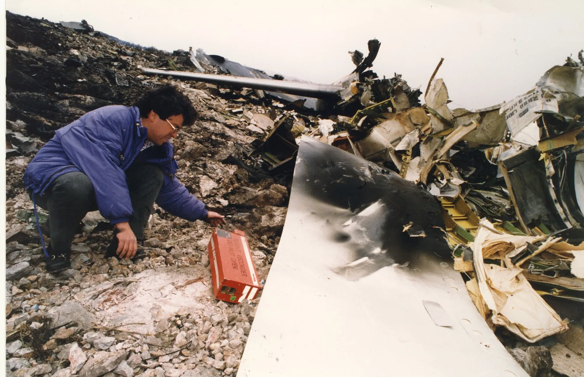 A man in a violet padded jacket crouches on a rocky slope, reaching towards an orange box with a white band on which «FLIGHT RECORDER — DO NOT OPEN» can be read. Around him extensive aircraft wreckage: a large white fuselage panel with a scorched area, twisted metal, insulation and a cylindrical strut. An overcast sky.