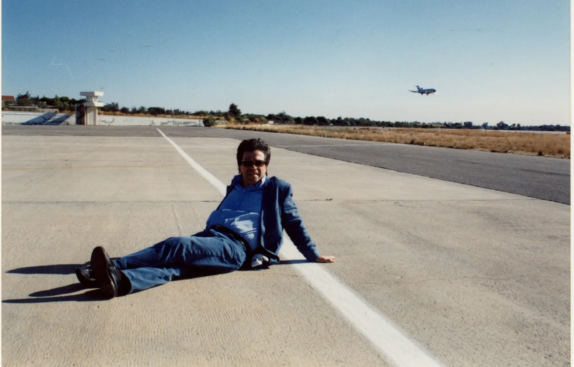 A man lies on his side on a concrete airport runway, propped on one elbow, in a denim jacket and jeans with sunglasses. The white centre line runs beside him. Far left, a tall glazed structure; at right, dry grass and trees; high and distant, an aircraft in flight. A clear blue sky.