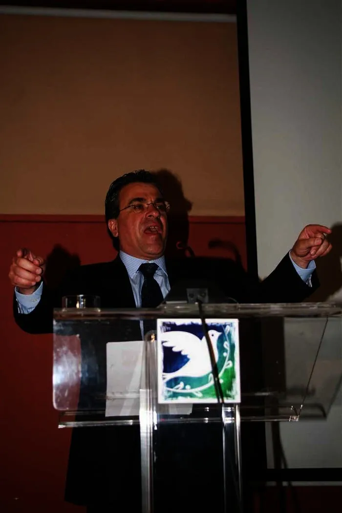 Ο Αργύρης Ντινόπουλος μιλά από το βήμα με υψωμένο χέρι.