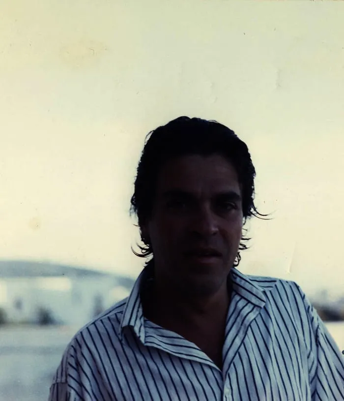 Κόντρα φωτισμένο πορτρέτο ενός άνδρα με ριγέ πουκάμισο μπροστά σε φωτεινό ουρανό και θάλασσα, σχεδόν σε σιλουέτα.