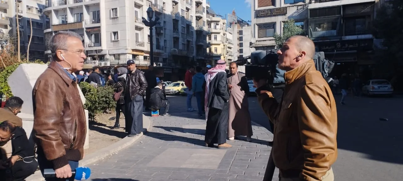 Ο Αργύρης Ντινόπουλος σε προφίλ αριστερά, με καφέ δερμάτινο μπουφάν και μικρόφωνο του ΑΝΤ1· απέναντί του εικονολήπτης με κάμερα στον ώμο. Ανάμεσά τους πλατεία της Δαμασκού με περαστικούς, πολυκατοικίες και ένα κίτρινο ταξί, Δαμασκός, Συρία, 2024.