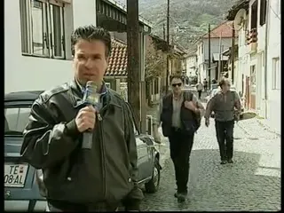 Ο Αργύρης Ντινόπουλος, με μαύρο δερμάτινο μπουφάν και μικρόφωνο στο χέρι, προχωρά προς τον φακό σε στενό λιθόστρωτο δρόμο με διώροφα σπίτια, κεραμοσκεπές και μπαλκόνια εκατέρωθεν· πίσω του περπατούν δύο περαστικοί και στα αριστερά είναι σταθμευμένο αυτοκίνητο με πινακίδα του Τετόβου· στο βάθος υψώνονται λόφοι.