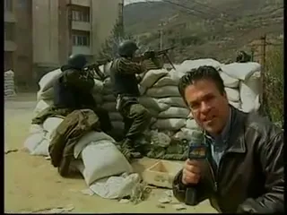 Ο Αργύρης Ντινόπουλος, με μαύρο δερμάτινο μπουφάν και μικρόφωνο στο χέρι, μιλά προς τον φακό στο δεξί μέρος του κάδρου· πίσω του, οδόφραγμα από στοιβαγμένα λευκά τσουβάλια με άμμο, όπου τέσσερις ένστολοι άνδρες με κράνη είναι σκυμμένοι, δύο από αυτούς σημαδεύοντας με τυφέκια πάνω από τα τσουβάλια· στο βάθος πολυκατοικίες και γυμνή πλαγιά λόφου, με συννεφιασμένο ουρανό.