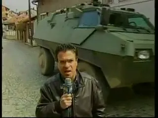 Ο Αργύρης Ντινόπουλος, με μαύρο δερμάτινο μπουφάν, στέκεται στο κέντρο λιθόστρωτου δρόμου κρατώντας μικρόφωνο και μιλά προς τον φακό· ακριβώς πίσω του, μεγάλο τροχοφόρο θωρακισμένο όχημα σε λαδί χρώμα, με πυργίσκο, καταλαμβάνει το πλάτος του δρόμου· εκατέρωθεν χαμηλά σπίτια με κεραμοσκεπές.
