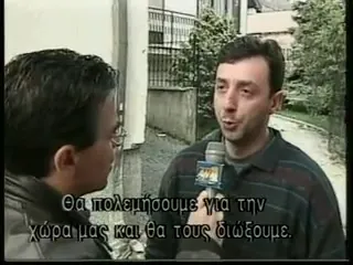 Νεαρός άνδρας με σκούρο μπουφάν μιλά σε συνέντευξη στο δεξί μέρος του κάδρου, κρατώντας μικρόφωνο με σημαιάκι τηλεοπτικού σταθμού· στα αριστερά, σε πρώτο πλάνο και σε προφίλ, το κεφάλι και ο ώμος του δημοσιογράφου που τον ακούει· πίσω τους λευκός τοίχος και βλάστηση. Στο κάτω μέρος του κάδρου, ενσωματωμένος τηλεοπτικός υπότιτλος της εκπομπής — «Θα πολεμήσουμε για την χώρα μας και θα τους διώξουμε.»