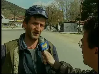 Ηλικιωμένος άνδρας με σκούρο τζόκεϊ, ανοιχτόχρωμο πουκάμισο και γκρι αμάνικο γιλέκο μιλά σε συνέντευξη στο κέντρο του κάδρου· από τα δεξιά, άνδρας γυρισμένος με την πλάτη στον φακό κρατά προς το μέρος του μικρόφωνο με σημαιάκι τηλεοπτικού σταθμού· πίσω τους άδειος δρόμος, χαμηλά κτίρια, γυμνά δέντρα και λόφοι.