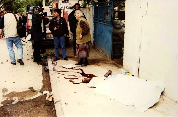 Σκηνή μετά από βομβαρδισμό: καλυμμένη σορός σε πεζοδρόμιο, ηλικιωμένη γυναίκα και συνεργείο Τύπου στο βάθος, Νις, 1999.
