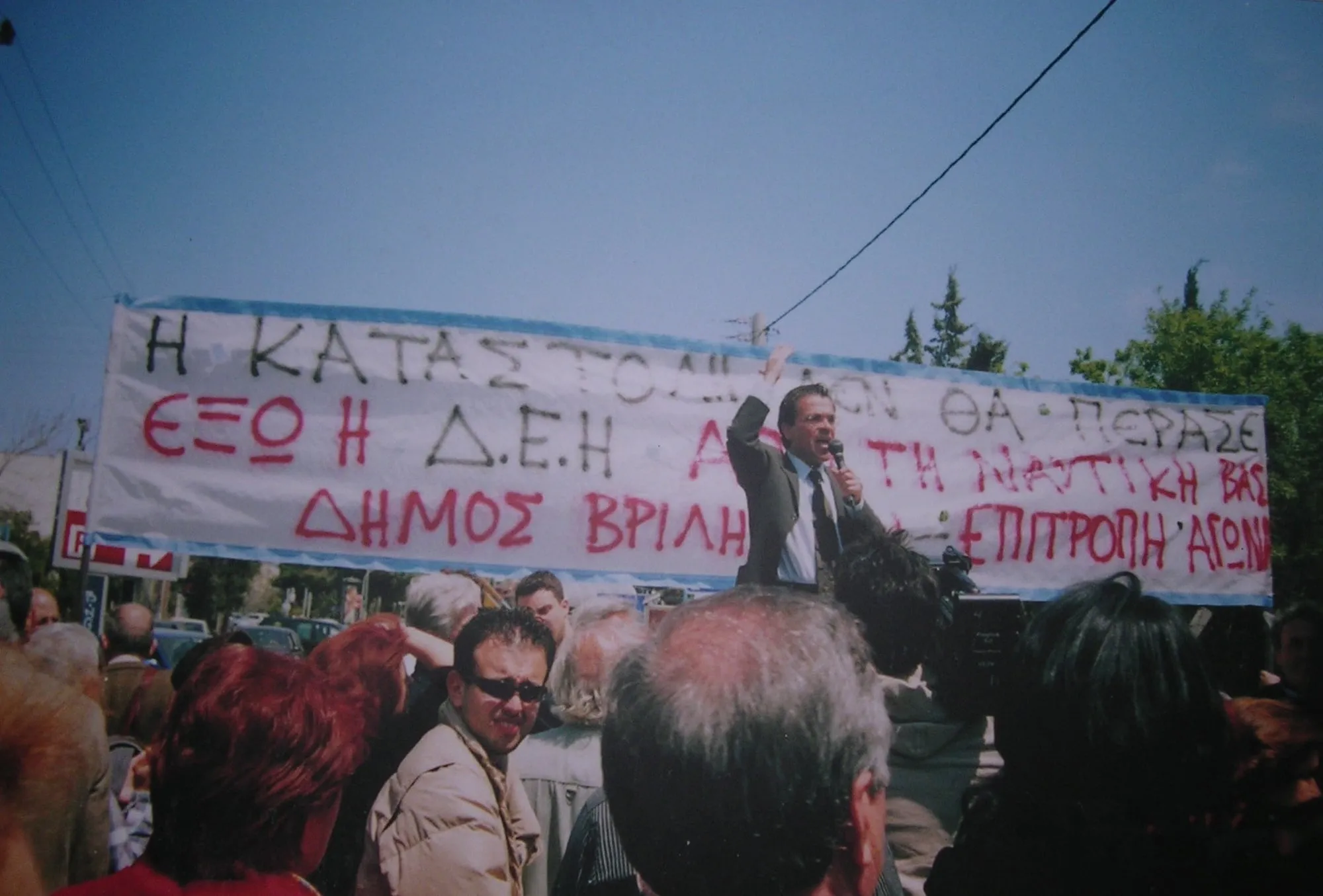 Ο Δήμαρχος Βριλησσίων Αργύρης Ντινόπουλος μιλά σε βραδινή συγκέντρωση μπροστά από μεγάλο χειροποίητο πανό διαμαρτυρίας.