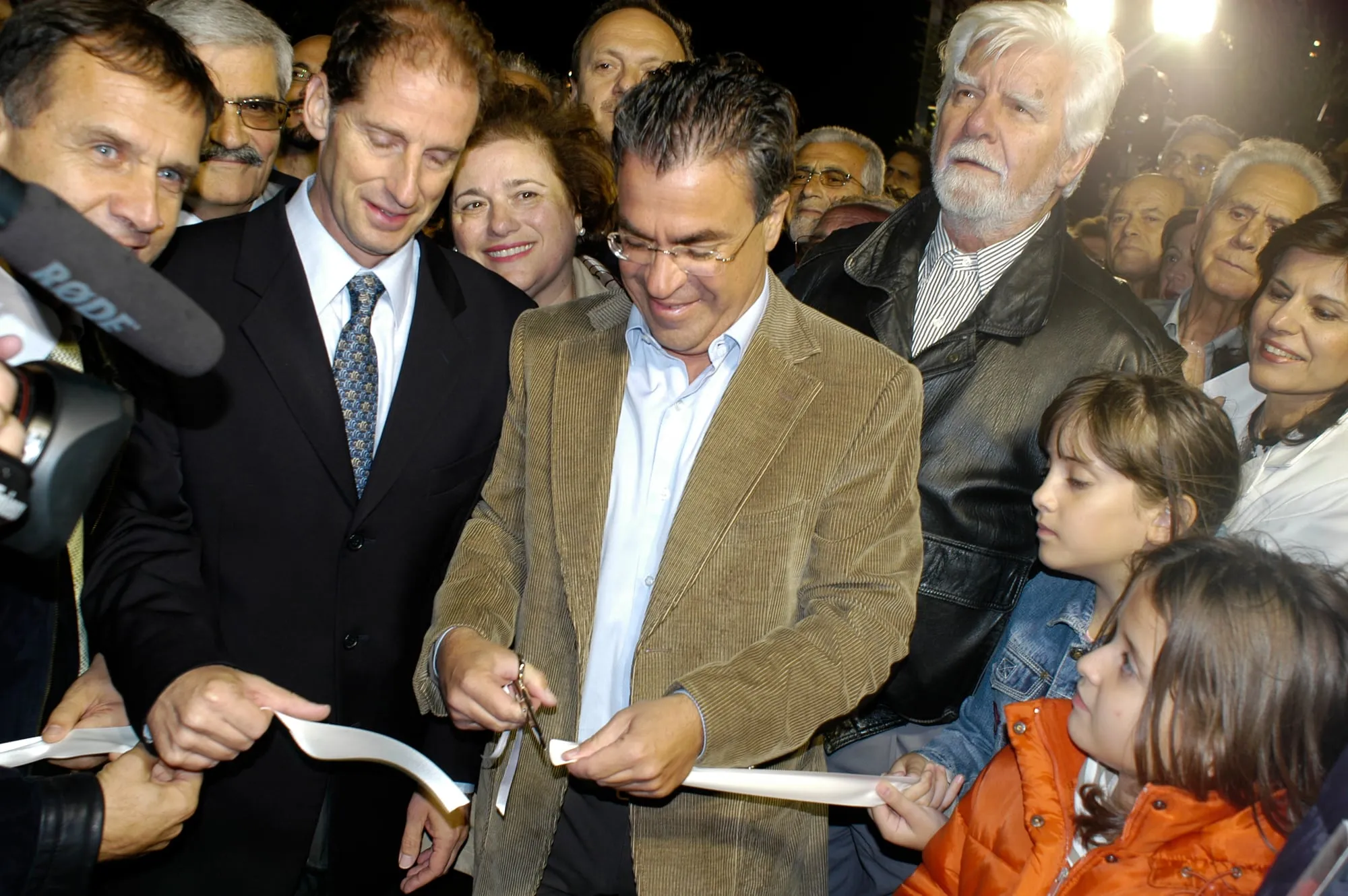 Βραδινή κοπή λευκής κορδέλας: ο Αργύρης Ντινόπουλος, με καφέ κοτλέ σακάκι και γυαλιά, κόβει την κορδέλα με ψαλίδι χαμηλώνοντας το βλέμμα, δίπλα του ψηλότερος άνδρας με σκούρο κοστούμι κρατά την άλλη άκρη. Δεξιά, στο πρώτο πλάνο, δύο κορίτσια — το ένα με τζιν τζάκετ, το άλλο με πορτοκαλί αμάνικο μπουφάν — κρατούν κι εκείνα την κορδέλα. Γύρω πυκνό πλήθος κατοίκων, μικρόφωνο τύπου και φώτα στα αριστερά.