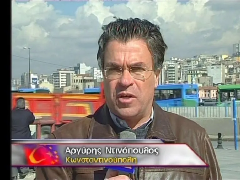 Καρέ τηλεοπτικής μετάδοσης — κοντινό πλάνο του Αργύρη Ντινόπουλου με δερμάτινο μπουφάν, να μιλά στην κάμερα κρατώντας μικρόφωνο· πίσω του δρόμος με λεωφορεία και η πυκνοδομημένη πόλη σε λόφο· κάτω αριστερά τηλεοπτικό πλαίσιο με σήμα τουρκικής σημαίας και αστεριών της ΕΕ και τις λέξεις «Αργύρης Ντινόπουλος / Κωνσταντινούπολη».