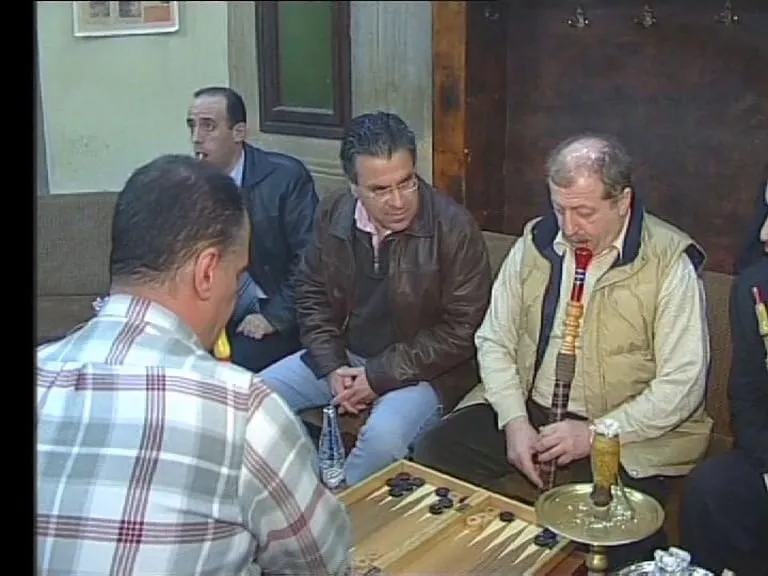Καρέ τηλεοπτικής μετάδοσης — σε καφενείο, άνδρες γύρω από χαμηλό τραπέζι· στο πρώτο πλάνο ταβλιέρα με τα πούλια της, στο κέντρο ο Αργύρης Ντινόπουλος με δερμάτινο μπουφάν καθισμένος ανάμεσά τους, και στα δεξιά ένας άνδρας, με σκυμμένο κεφάλι, φέρνει στα χείλη του το επιστόμιο ενός ναργιλέ που στέκεται σε κίτρινο μεταλλικό δίσκο.