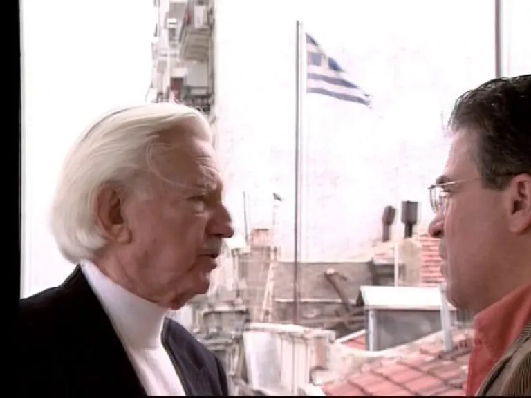 Καρέ τηλεοπτικής μετάδοσης — ηλικιωμένος άνδρας με λευκά μαλλιά και μουστάκι, σε προφίλ, μιλά στον Αργύρη Ντινόπουλο, απέναντί του· πίσω τους, σε φωτεινό φόντο, κεραμοσκεπές με καμινάδες και μια ελληνική σημαία που κυματίζει σε ιστό.