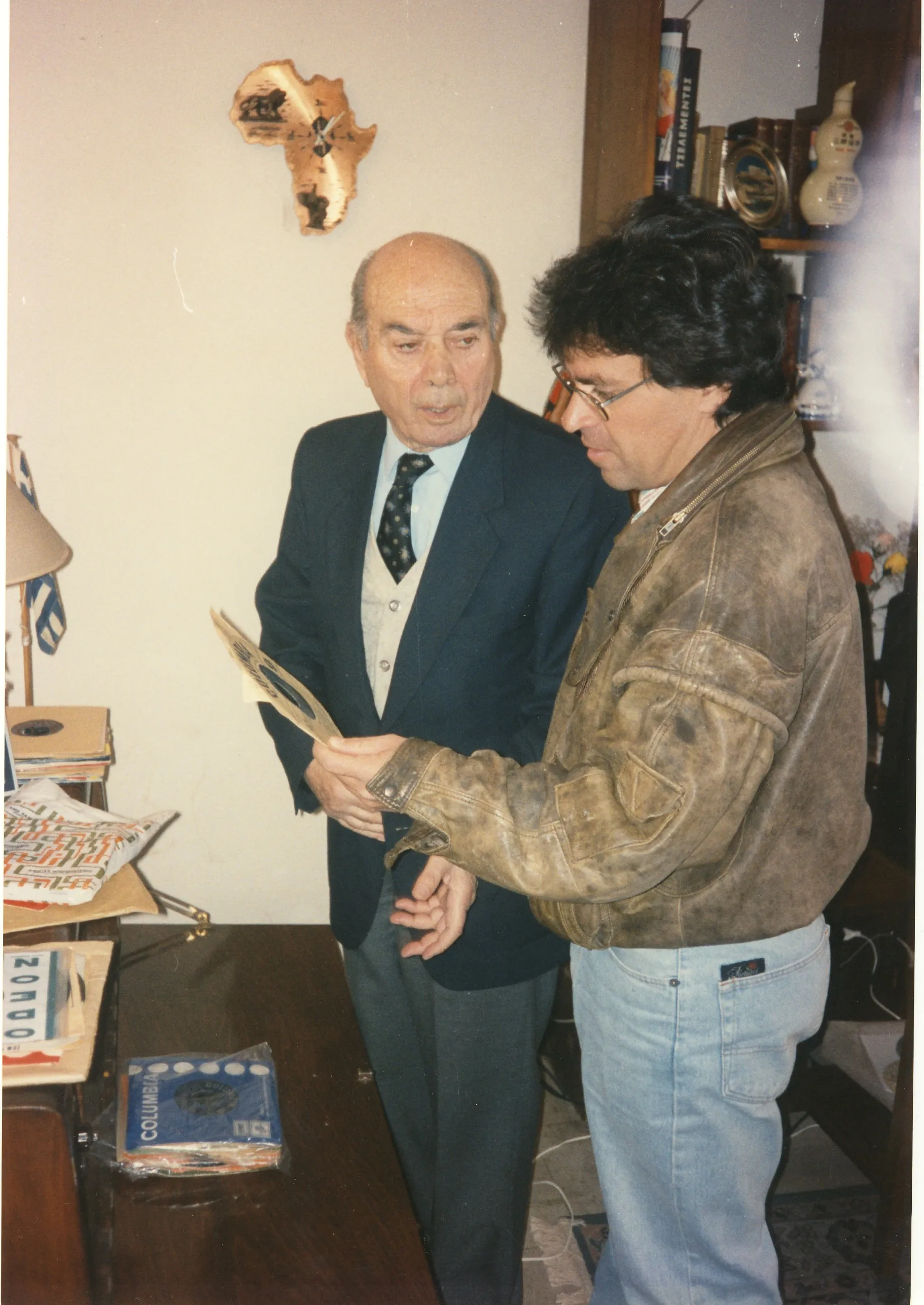 Indoors, an elderly man in a dark suit, pale knitted waistcoat and patterned tie holds a record in a paper sleeve and is speaking; beside him Argyris Dinopoulos, in glasses and a worn leather jacket, leans in to look at it; at the upper left a clock in the shape of Africa hangs on the wall, to the right a shelf of books and mementoes, and on a wooden table in front of them stacks of records with ODEON and COLUMBIA sleeves; at the left edge a small Greek flag is visible.