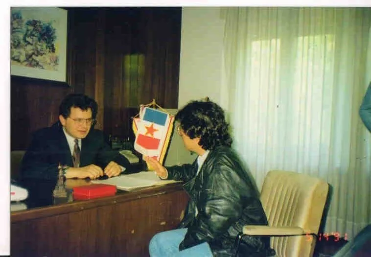 Ο Αργύρης Ντινόπουλος, με την πλάτη στον φακό, παίρνει συνέντευξη από τον Μίλαν Μπάμπιτς σε ξύλινο γραφείο, με ένα μικρό γιουγκοσλαβικό σημαιάκι στημένο ανάμεσά τους, Κνιν, Απρίλιος 1991.