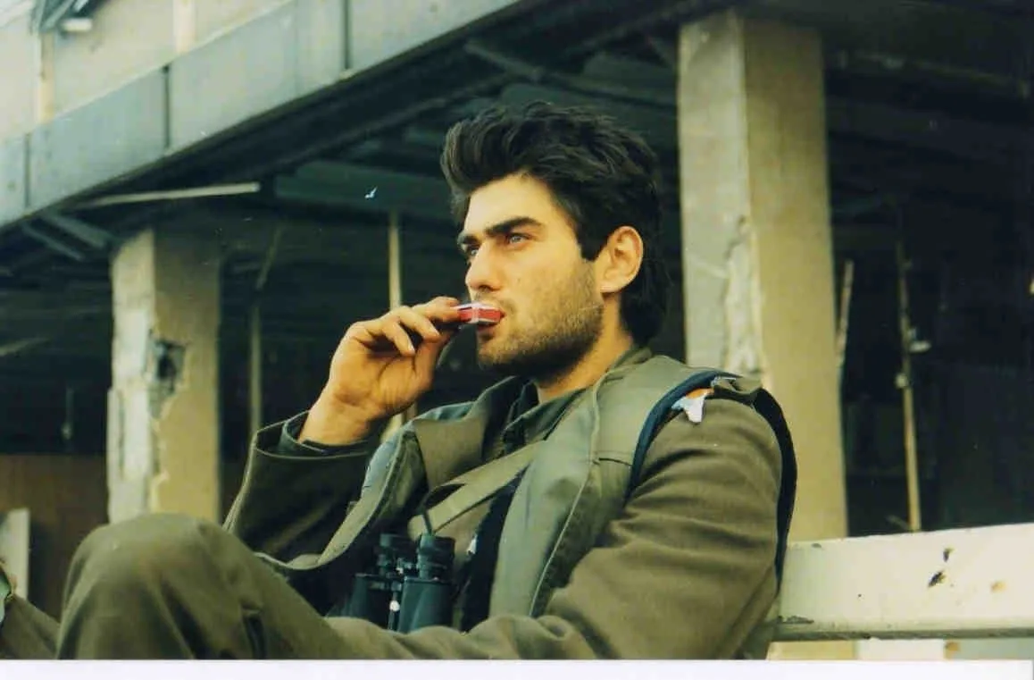Νεαρός άνδρας με στρατιωτικό χιτώνιο και κιάλια στον λαιμό, καθισμένος μπροστά σε κατεστραμμένο κτίριο, φέρνει στα χείλη του ένα πακέτο τσιγάρα, Βούκοβαρ, 1991.