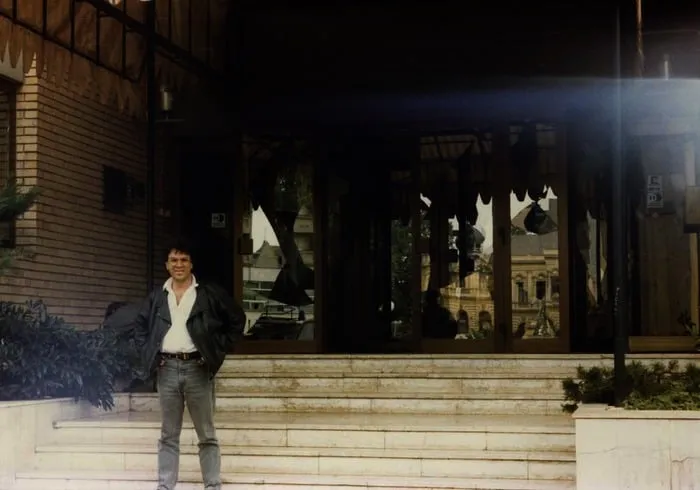 Ο Αργύρης Ντινόπουλος μπροστά σε κτίριο με σπασμένες βιτρίνες, Βούκοβαρ, 1991.
